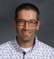 A picture of Tim Davis, award winning teacher, in a white button down shirt with a purple pattern against a gray backdrop 