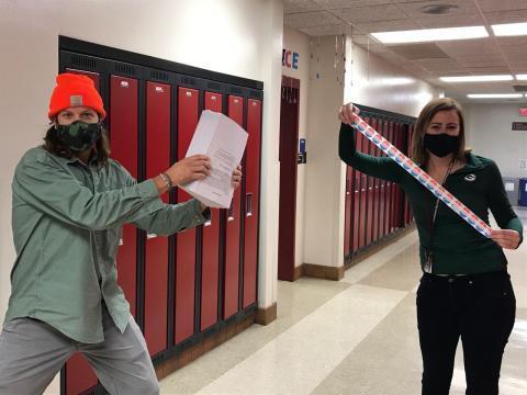 A teacher holds up I voted stickers while another teacher holds ballots. Both teachers are wearing masks due to Covid-19. They are in the school hallway six feet apart. 
